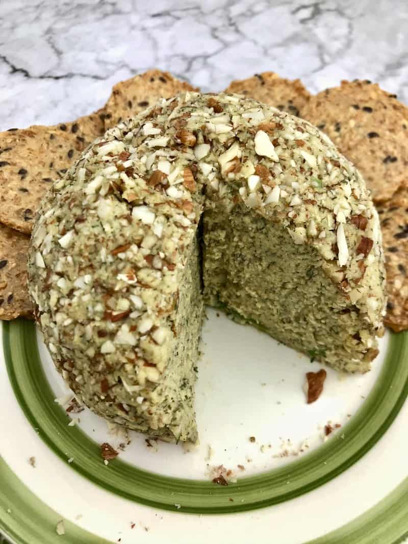 A cashew cheese ball with a section cut out, sitting on a plate next to round crackers.