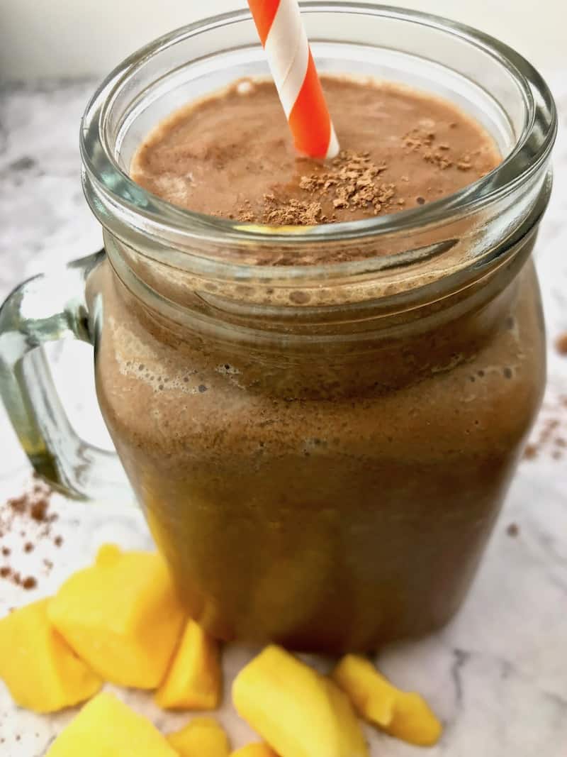 A chocolate smoothie with an orange and white straw, sitting next to mango chunks.