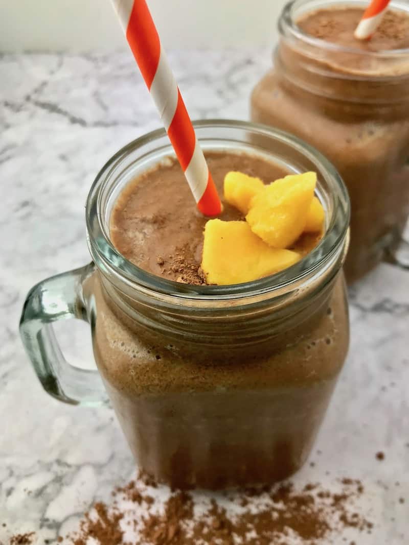 A chocolate smoothie with mango pieces on top of it and an orange and white straw in it.