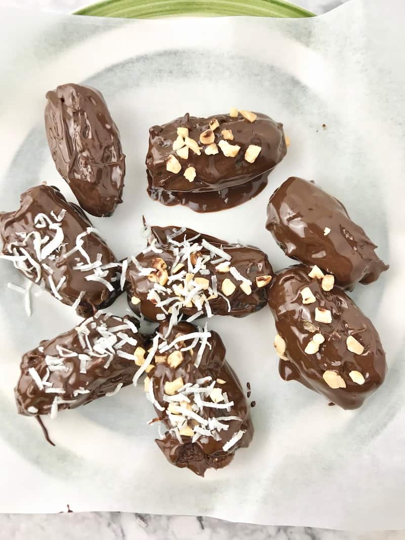 Chocolate-dipped dates covered in coconut flakes and chopped peanuts, sitting on a parchment-lined plate.