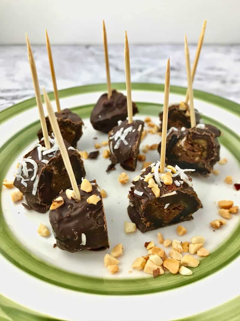 Pieces of chocolate covered dates stuck with toothpicks on a plate with chopped peanuts.