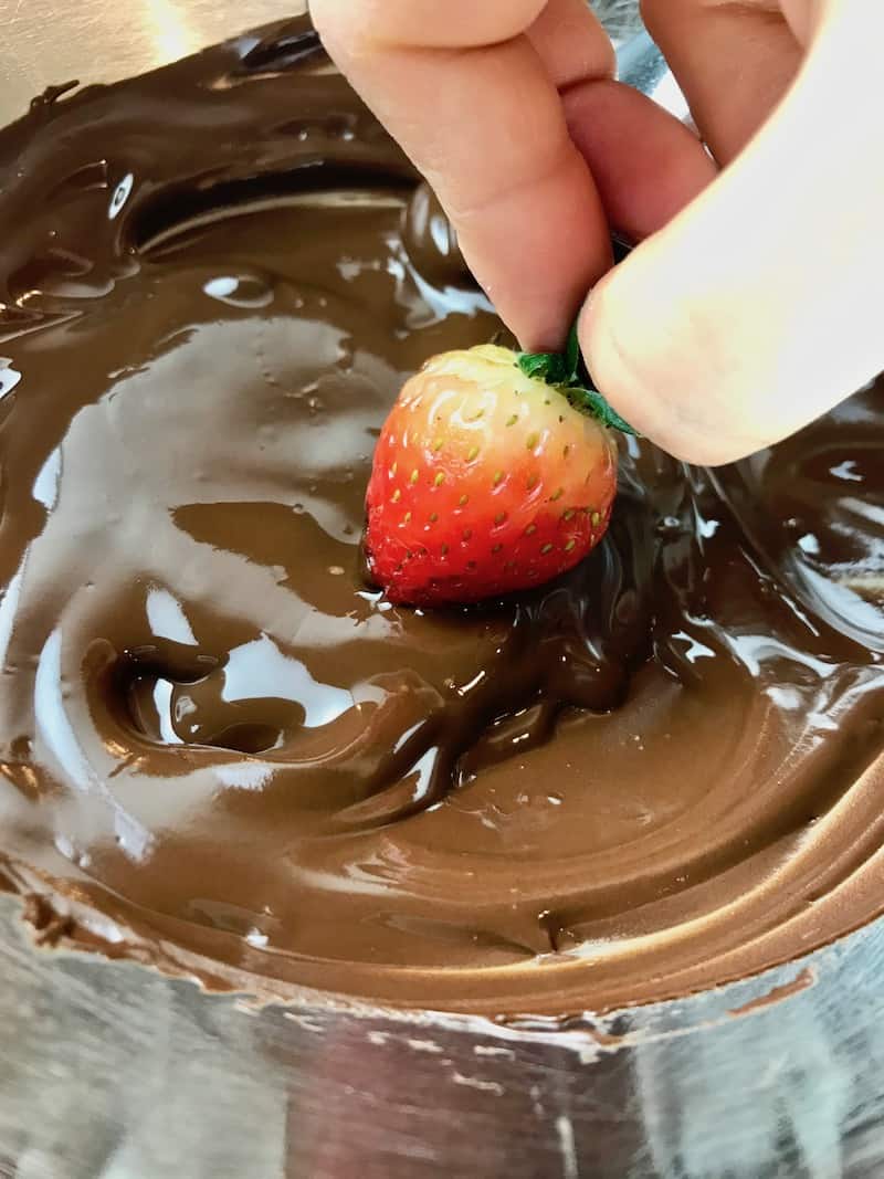 A hand dipping a strawberry in melted chocolate.