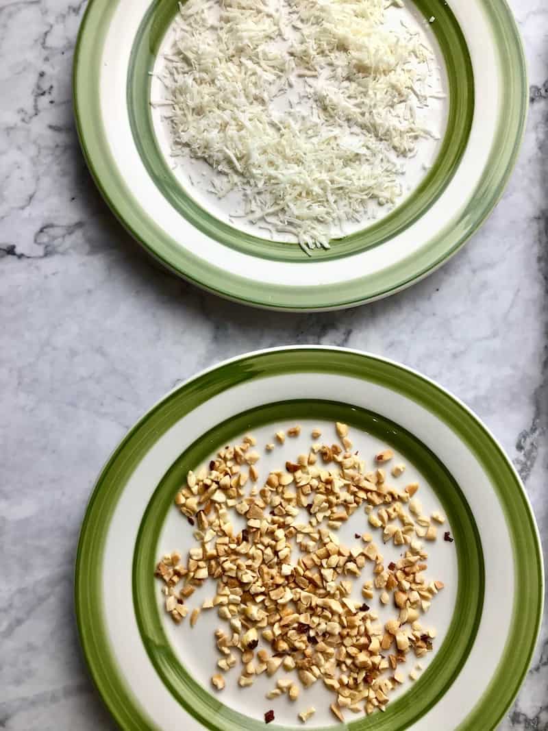 A plate of chopped peanuts next to a plate of coconut flakes.