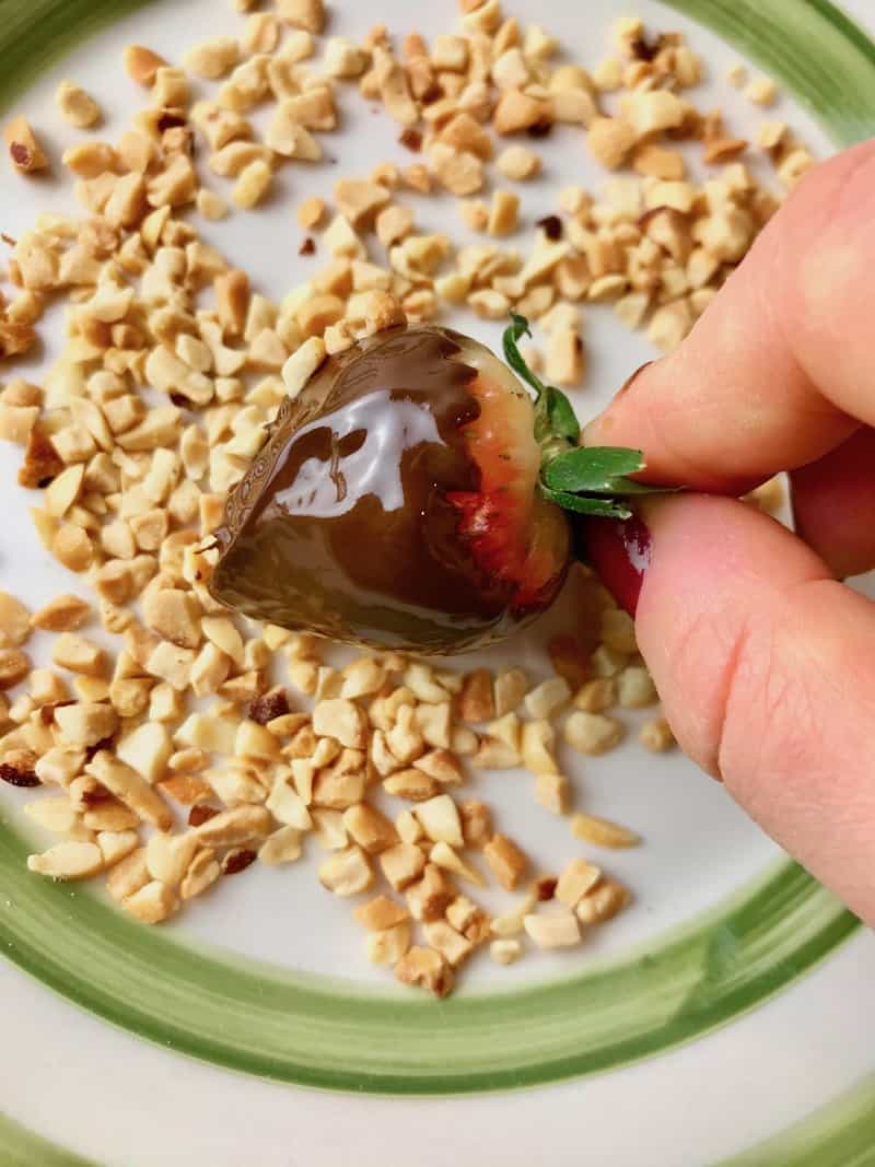 A hand rolling a chocolate dipped strawberry in chopped peanuts.