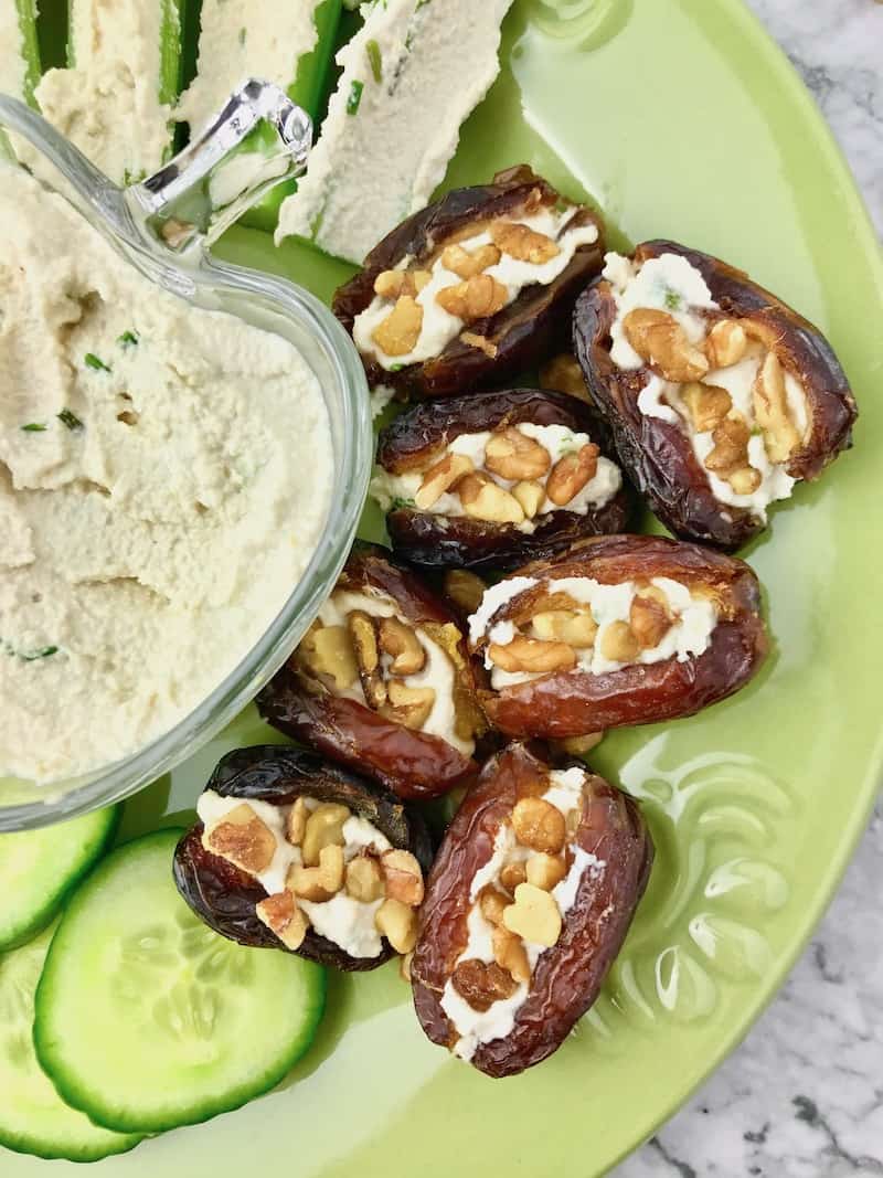 Stuffed dates topped with walnuts next to a bowl of cream cheese, celery sticks, and cucumber slices.