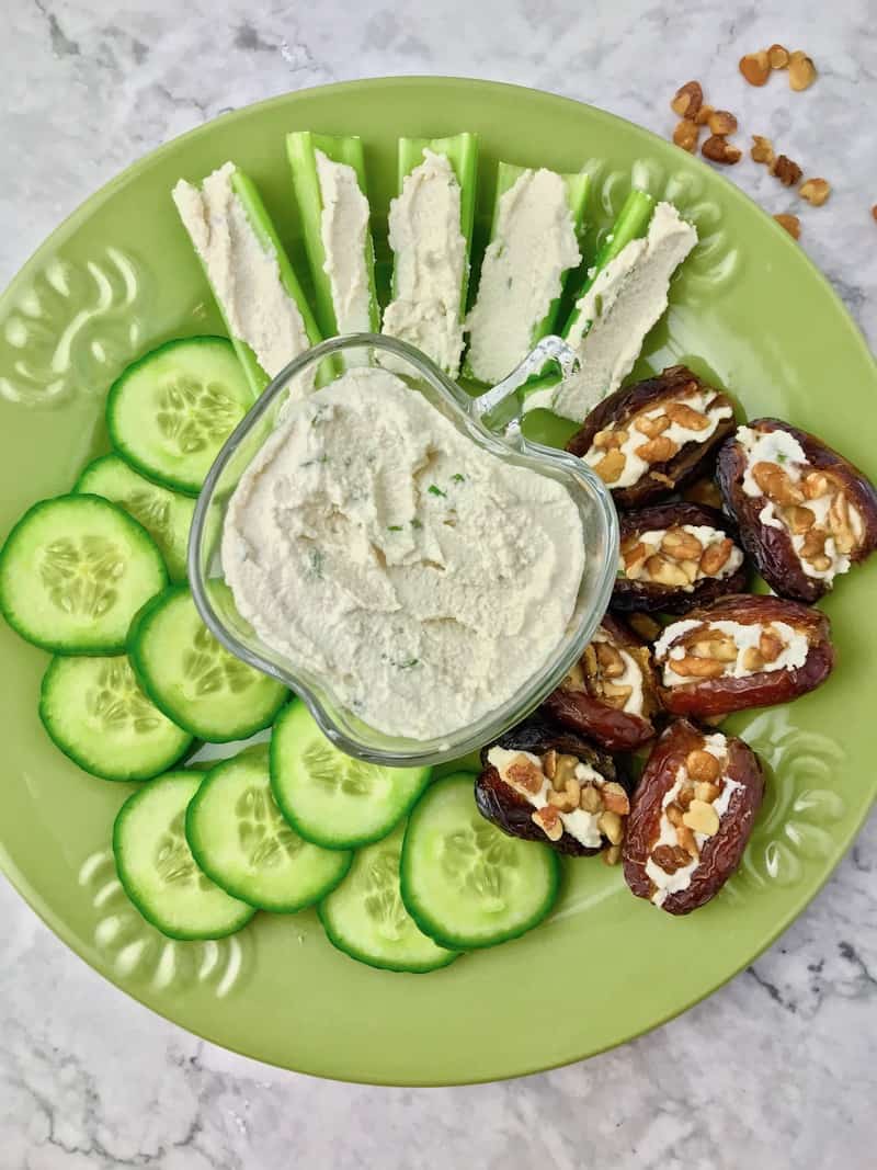 A plate with a bowl of vegan cream cheese surrounded by cucumber, celery, and stuffed dates.