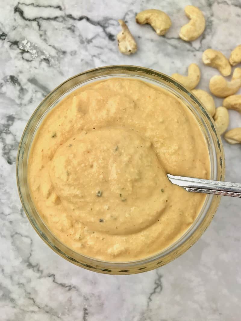 A small bowl of vegan burger sauce with a spoon, sitting next to some cashews.