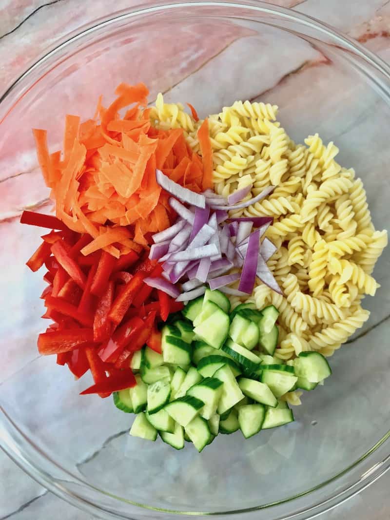 A bowl filled with pasta, carrot, onion, red pepper, and cucumber.