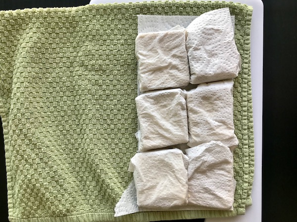 Slices of tofu wrapped in paper towel, sitting on a dish towel.