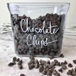 A container of chocolate chips with some chocolate chips scattered in front of it.