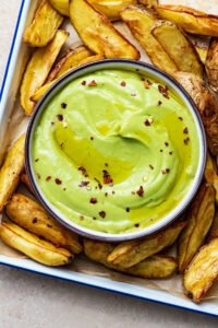 Avocado aioli in a small bowl surrounded by potato wedges.