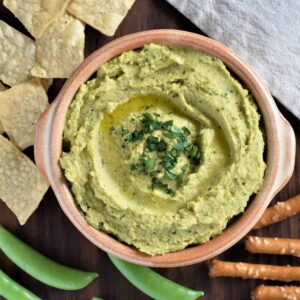 Avocado hummus in a bowl next to snap peas, pretzel sticks, and chips.