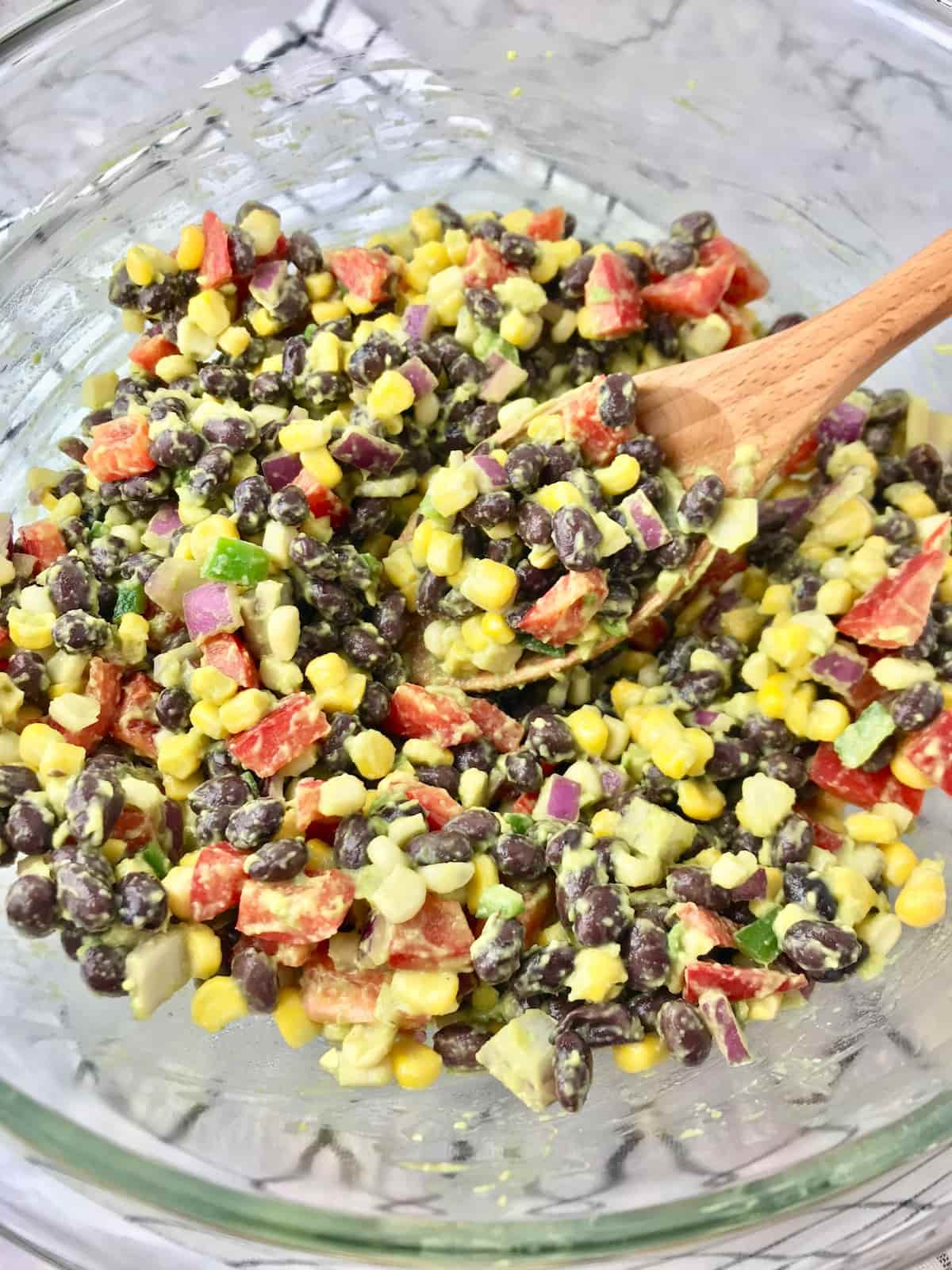 A large clear bowl full of black bean and corn salad with a wooden spoon.