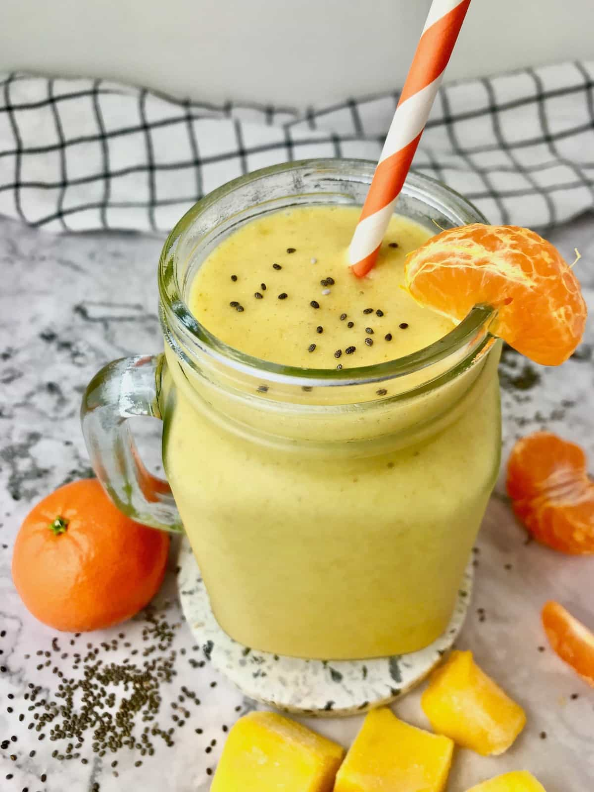 A yellow smoothie next to oranges and frozen mango chunks.