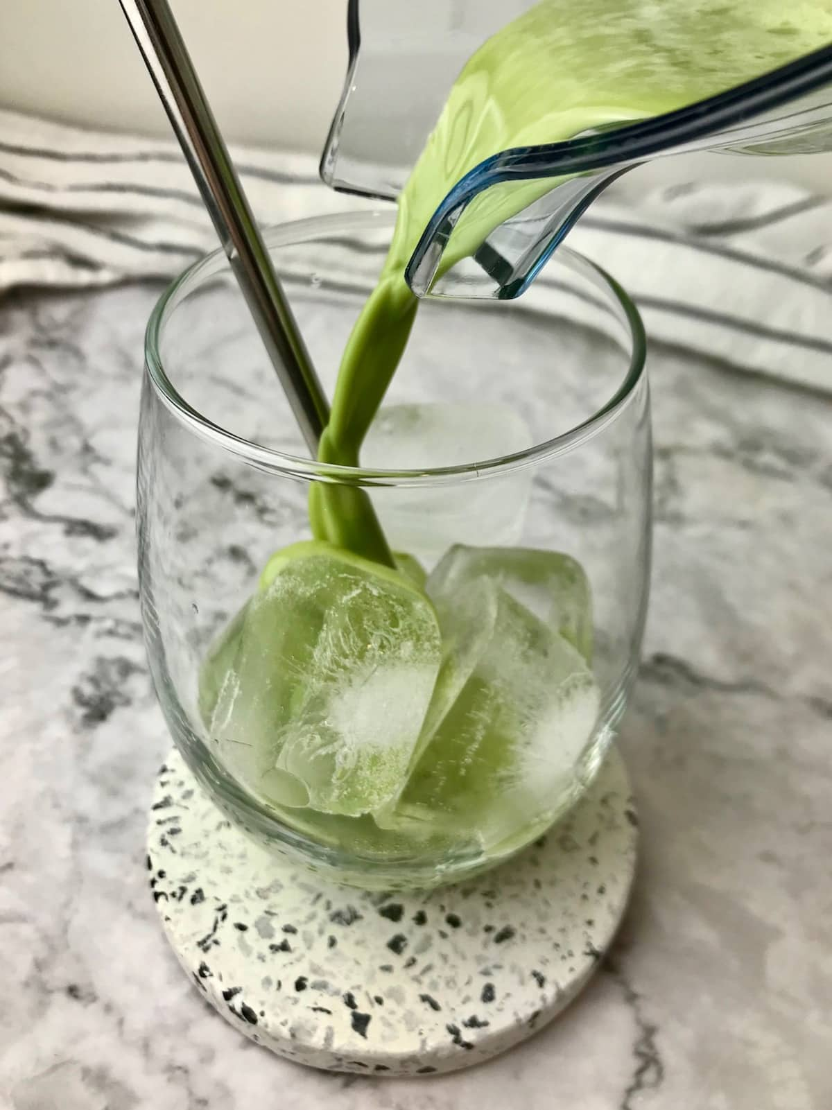 A match latte being poured into a stemless wine glass with ice cubes in it.