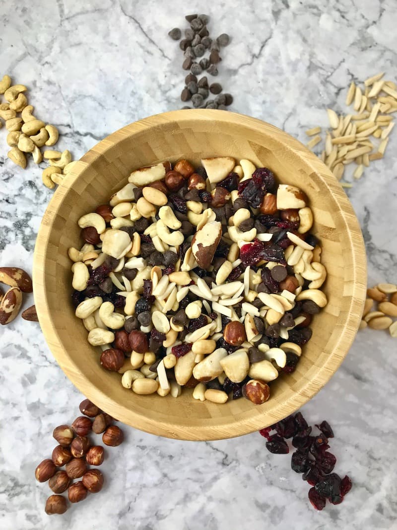 A wooden bowl of trail mix surrounded by various nuts, chocolate chips, and dried cranberries.