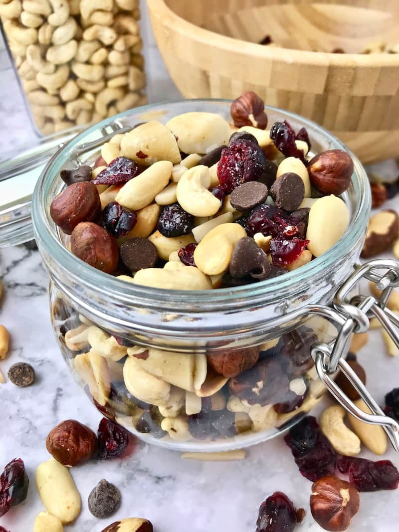 A glass jar of trail mix with some scattered in front of the jar.