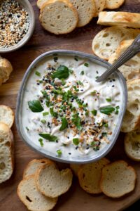 A white caramelized shallot dip topped with seasoning and herbs, surrounded by bread pieces.