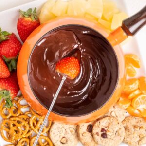Chocolate fondue with a strawberry dipped into it, surrounded by fruit, pretzels, and cookies.