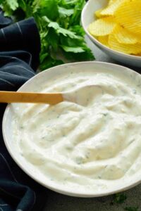 A bowl of vegan ranch dip with a wooden spoon in it, next to some potato chips.