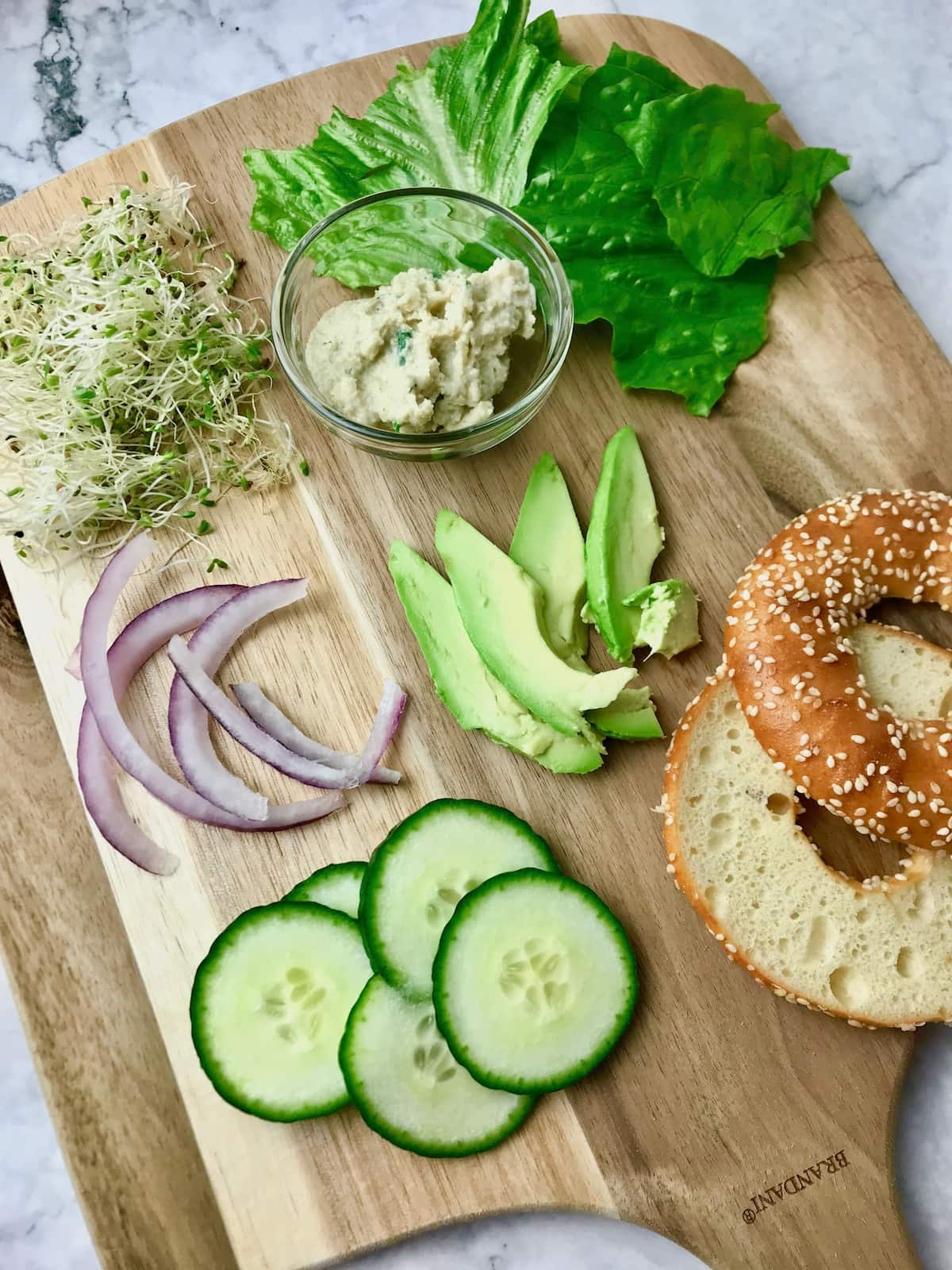 A tray of ingredients including lettuce, vegan cream cheese, onion, sprouts, cucumber, avocado, and a bagel.