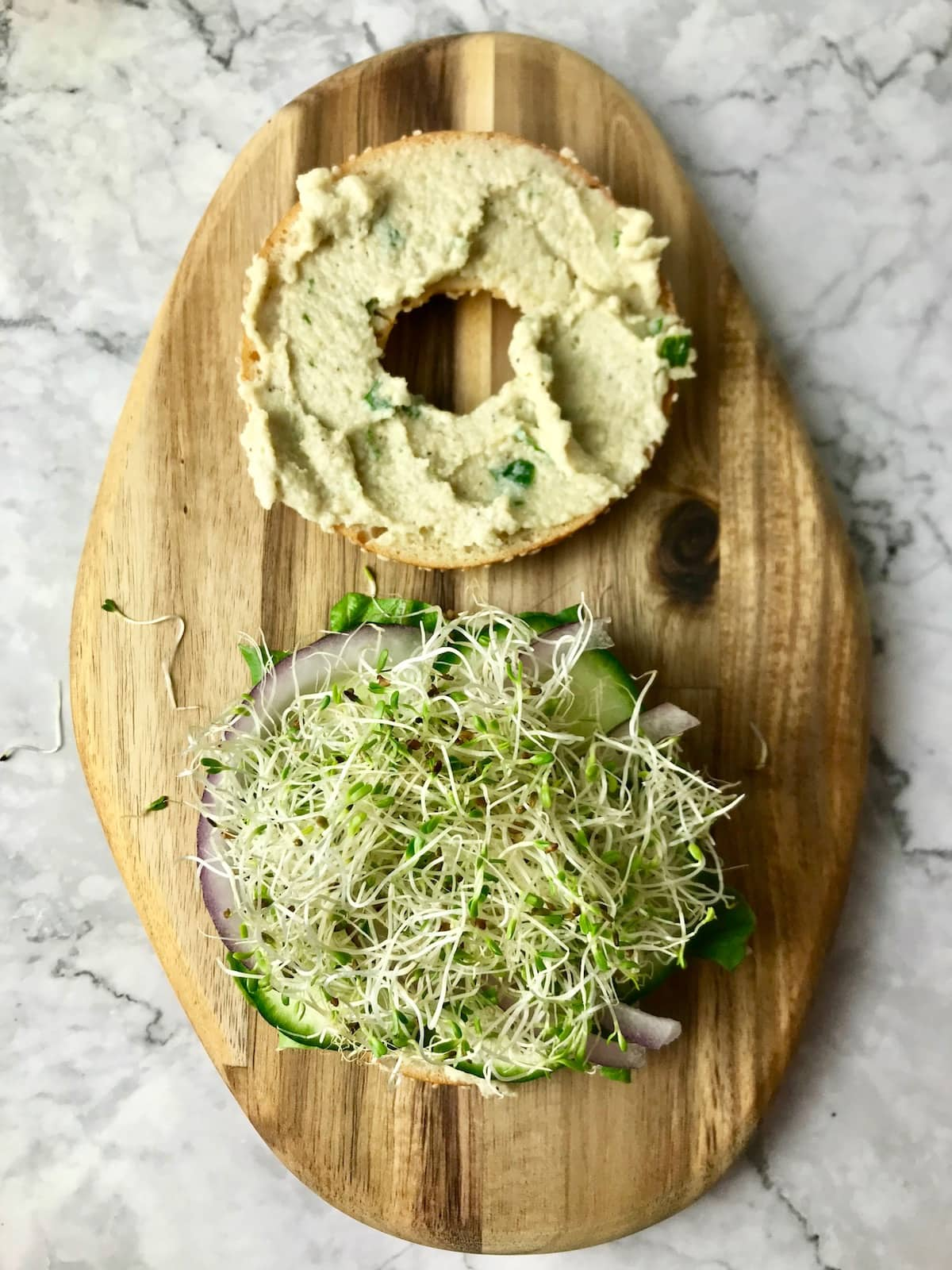 A bagel slice topped with cream cheese and another slice topped with veggies including onion, hummus, and sprouts..