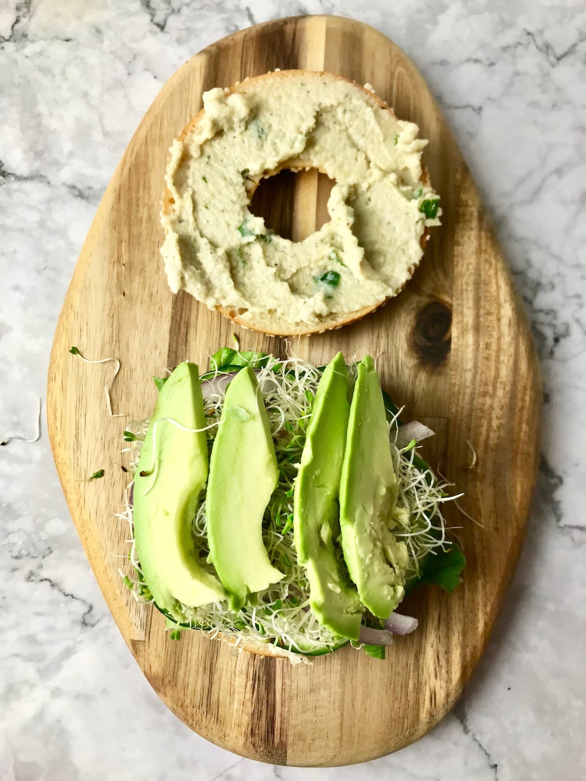 A bagel slice topped with cream cheese and another slice topped with onion, sprouts, and avocado.