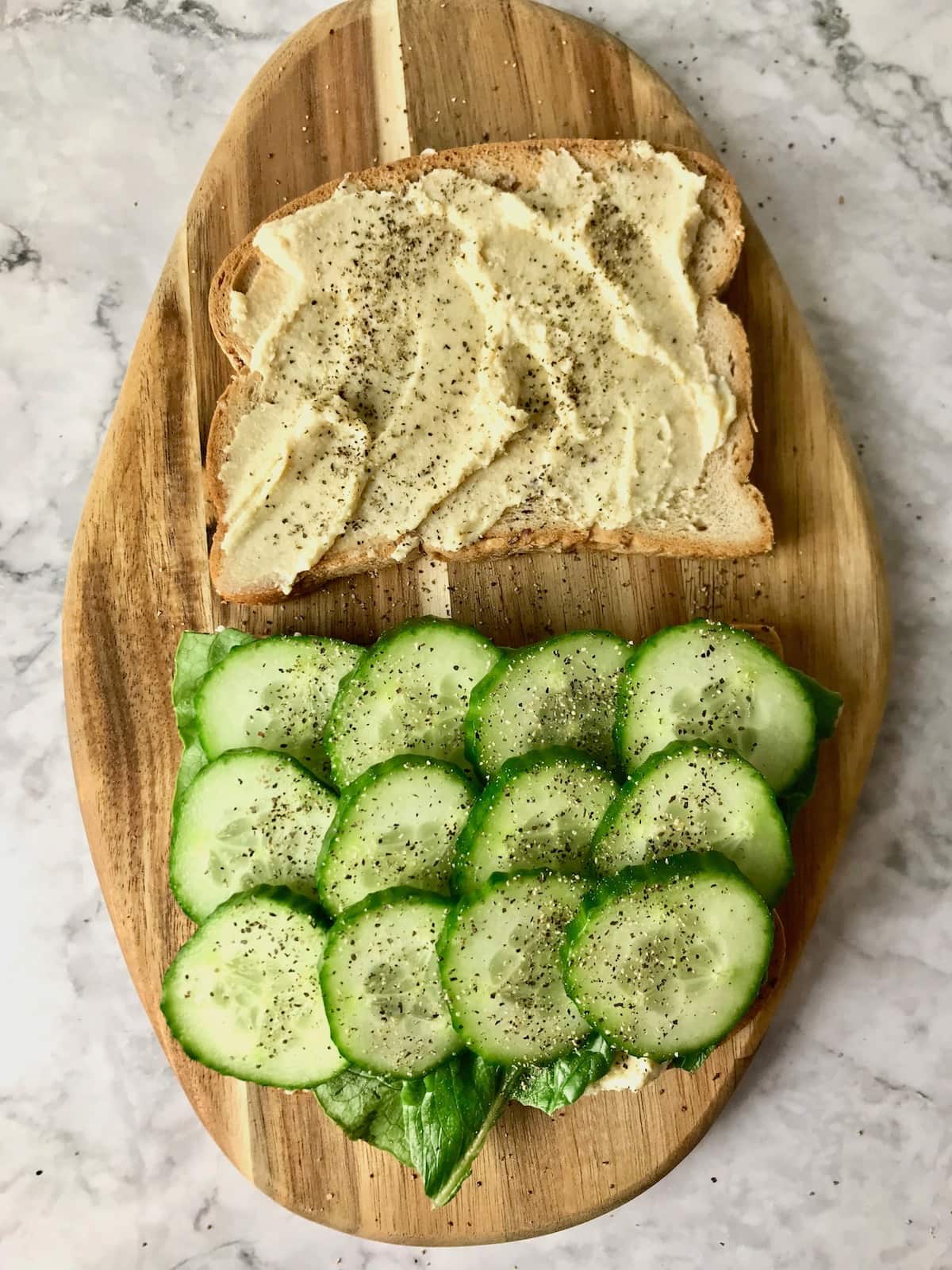 Two slices of bread, one with hummus, one with sliced cucumbers topped with black pepper.