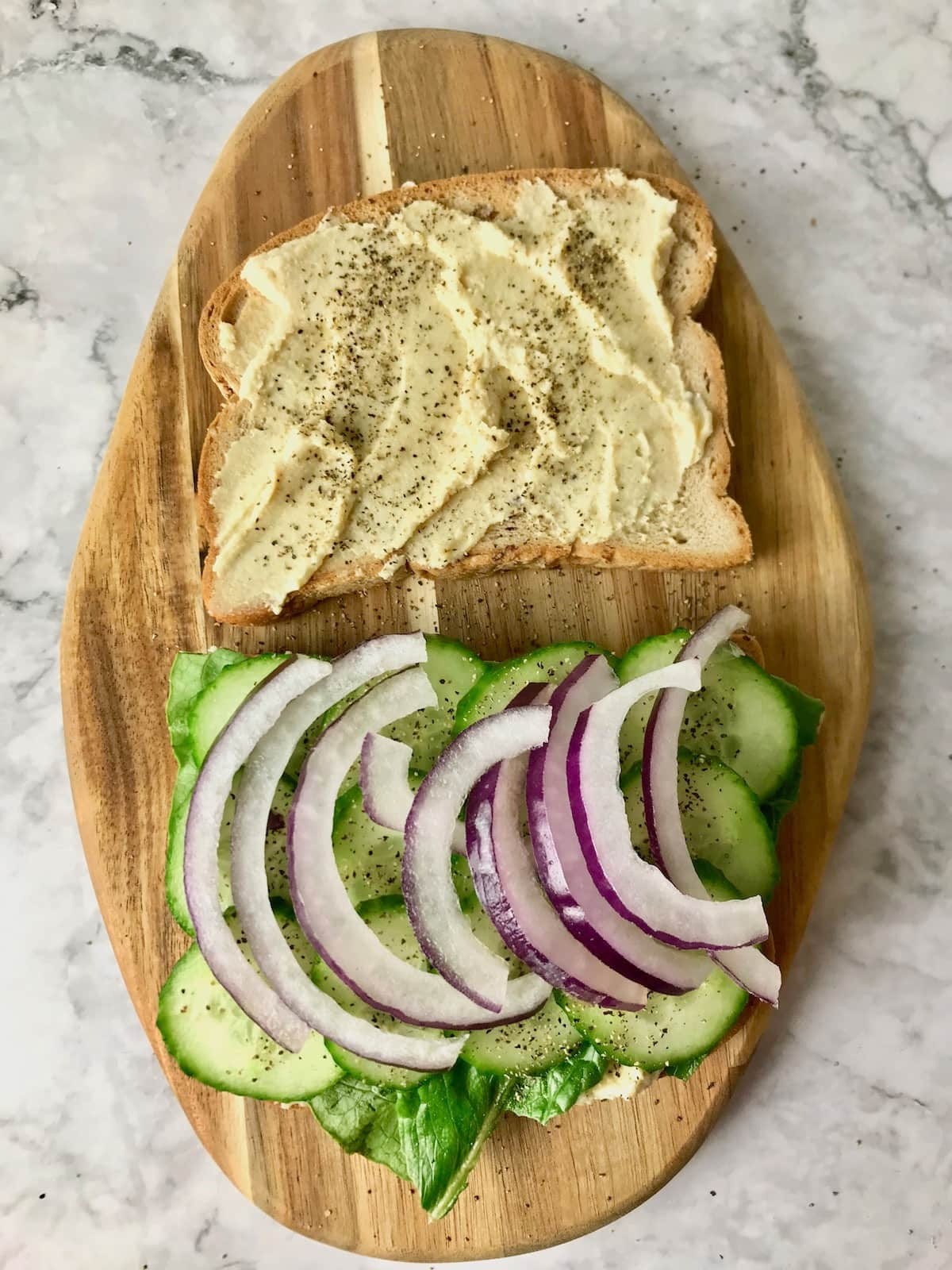 Two slices of bread, one topped with hummus, the other with cucumber and onion.