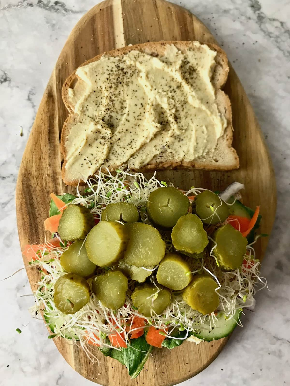 Two slices of bread, one topped with hummus, the other topped with various veggies, with pickles on top.