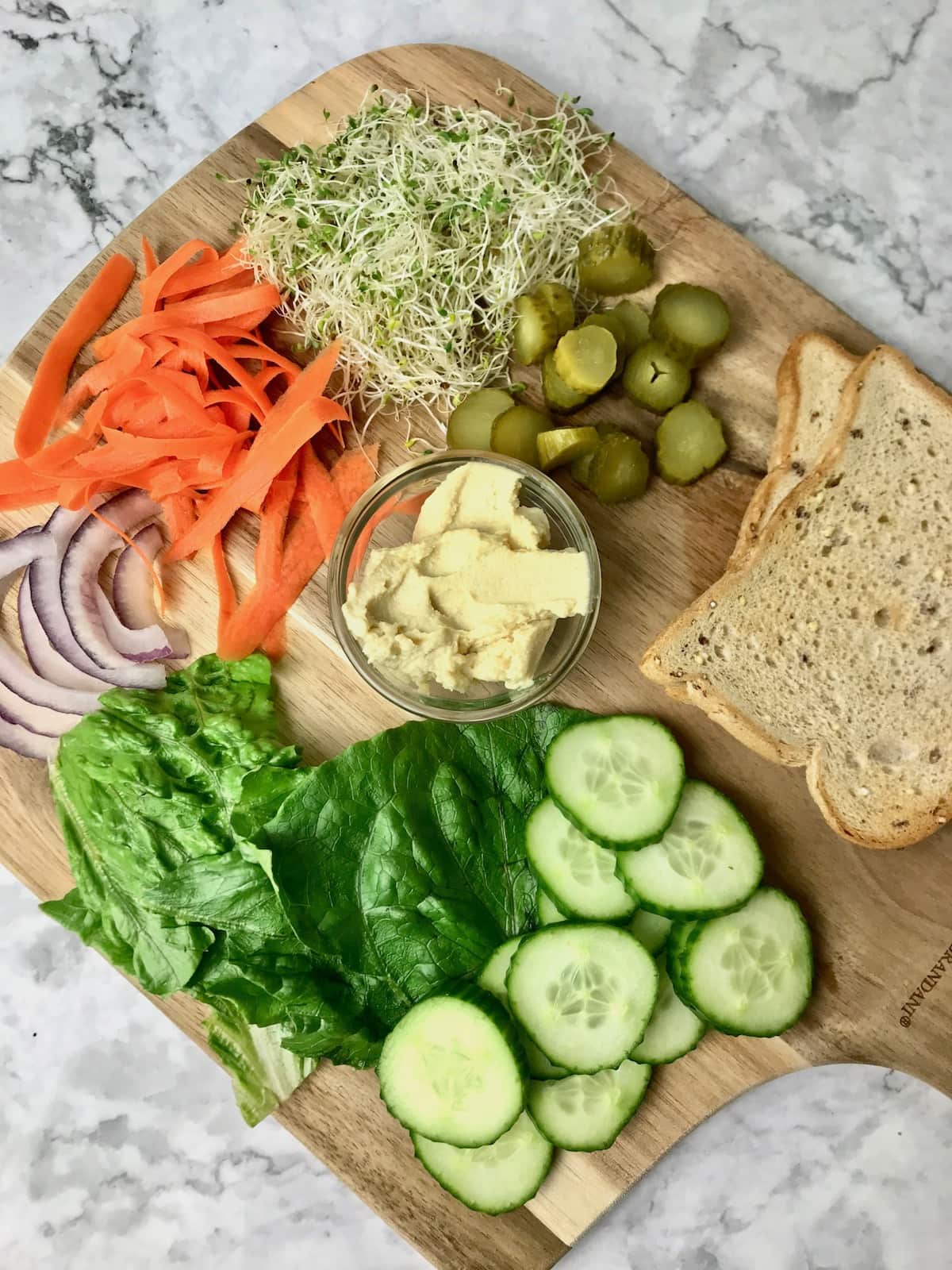 A board of sandwich ingredients, including hummus, pickles, cucumber, carrot, onion, sprouts, and lettuce.