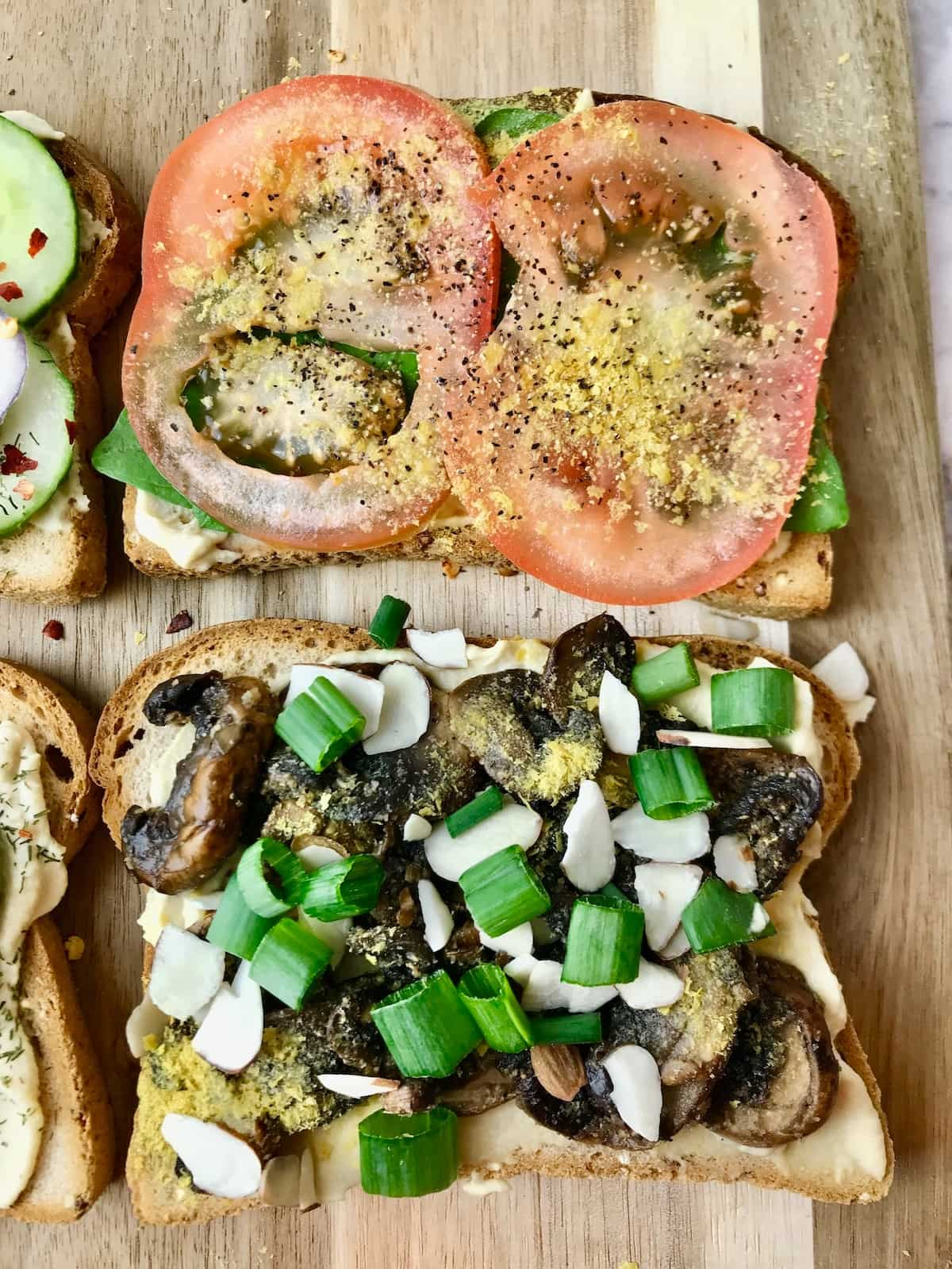 Two slices of hummus toast, one topped with sliced tomato, and the other topped with mushrooms.