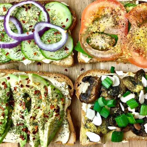 Four pieces of hummus toast, topped with cucumber, tomato, avocado, and mushrooms.