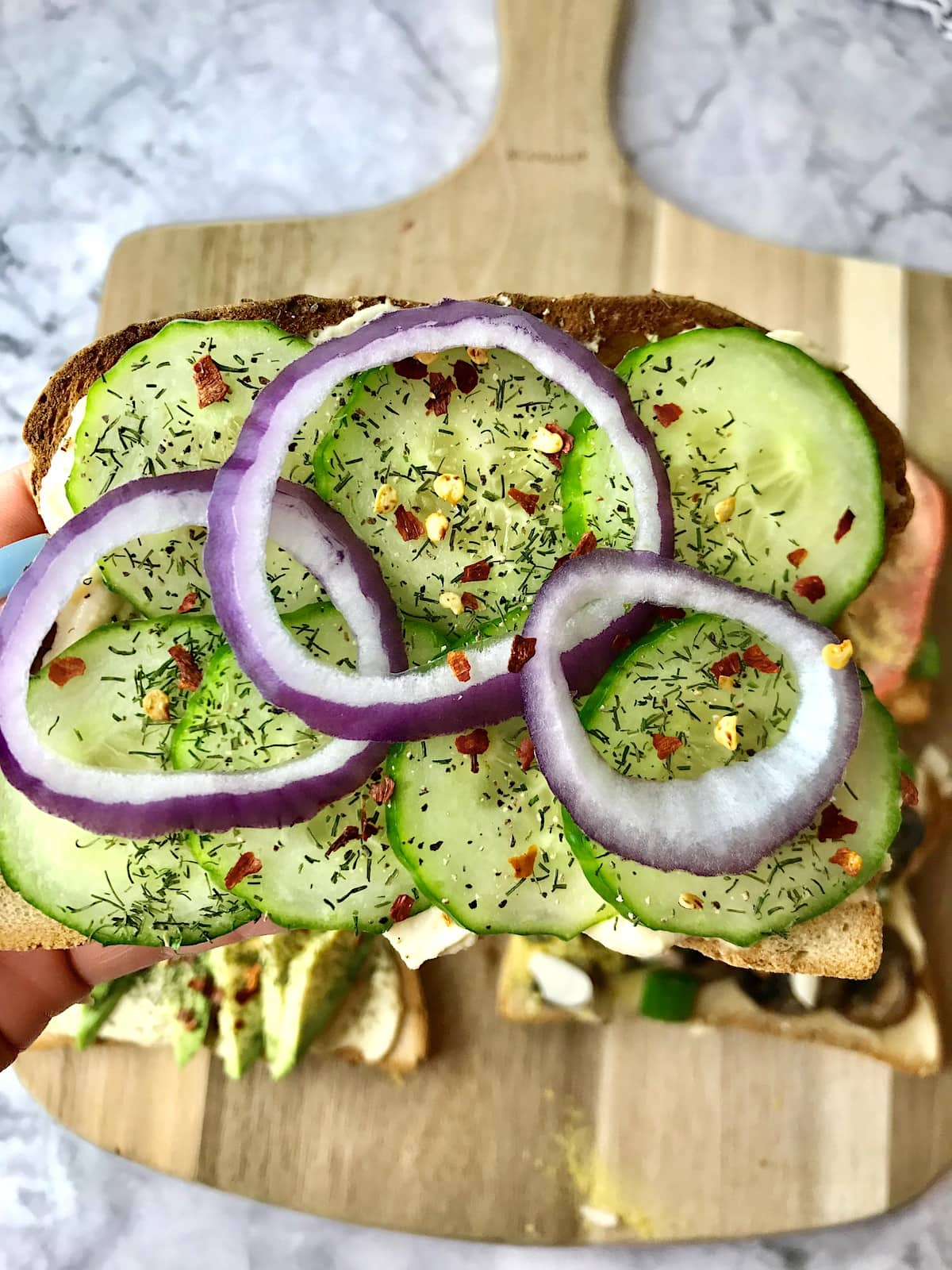 A piece of hummus toast topped with sliced cucumber and red onion.