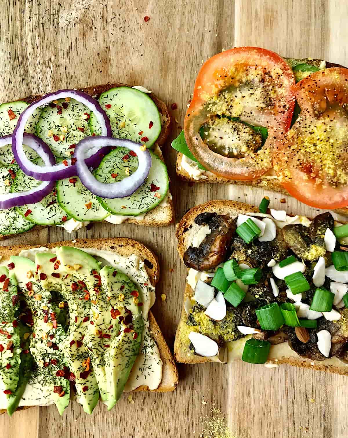Four slices of hummus toast with various toppings on a wooden cutting board.