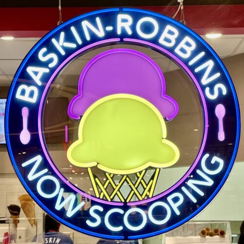 A lit up sign of an ice cream cone that says Baskin Robbins Now Scooping.
