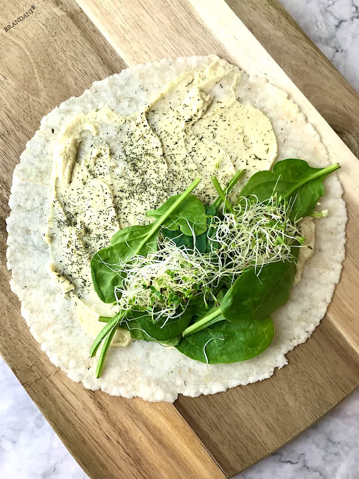 A tortilla topped with hummus, dill, spinach, and sprouts.