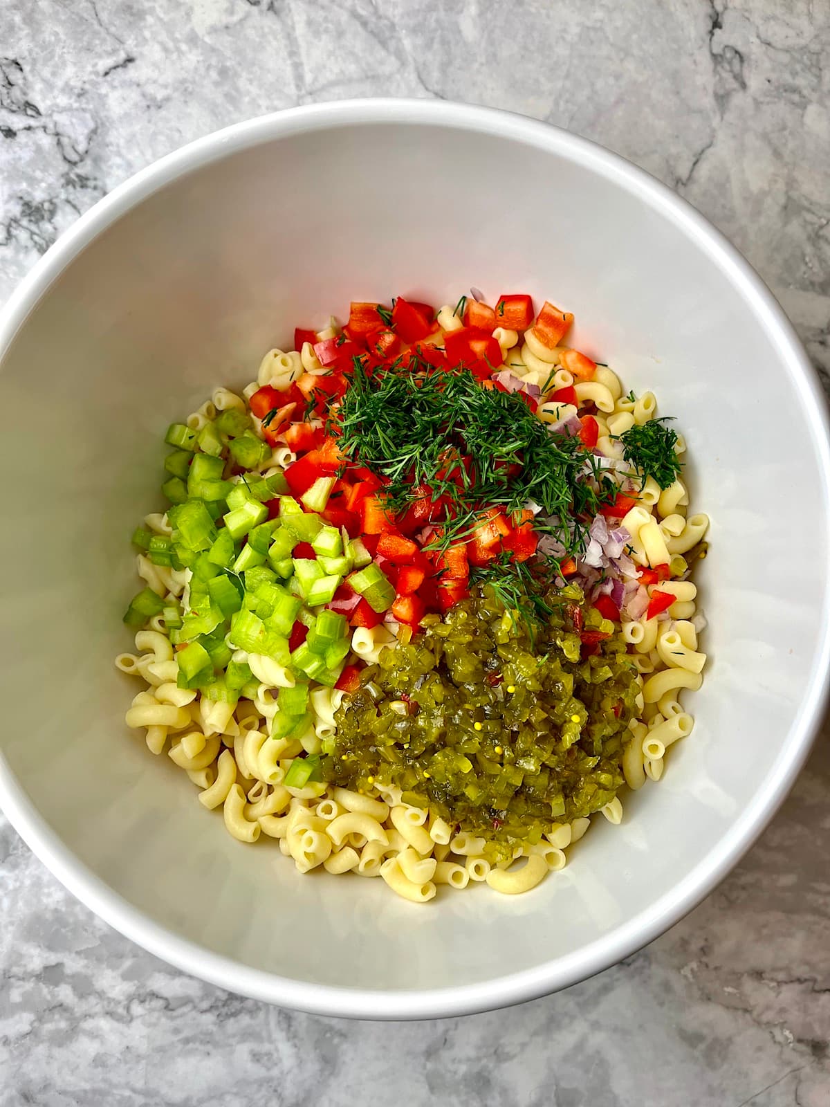 A bowl of macaroni salad ingredients including celery, red onion, red pepper, relish, and dill.