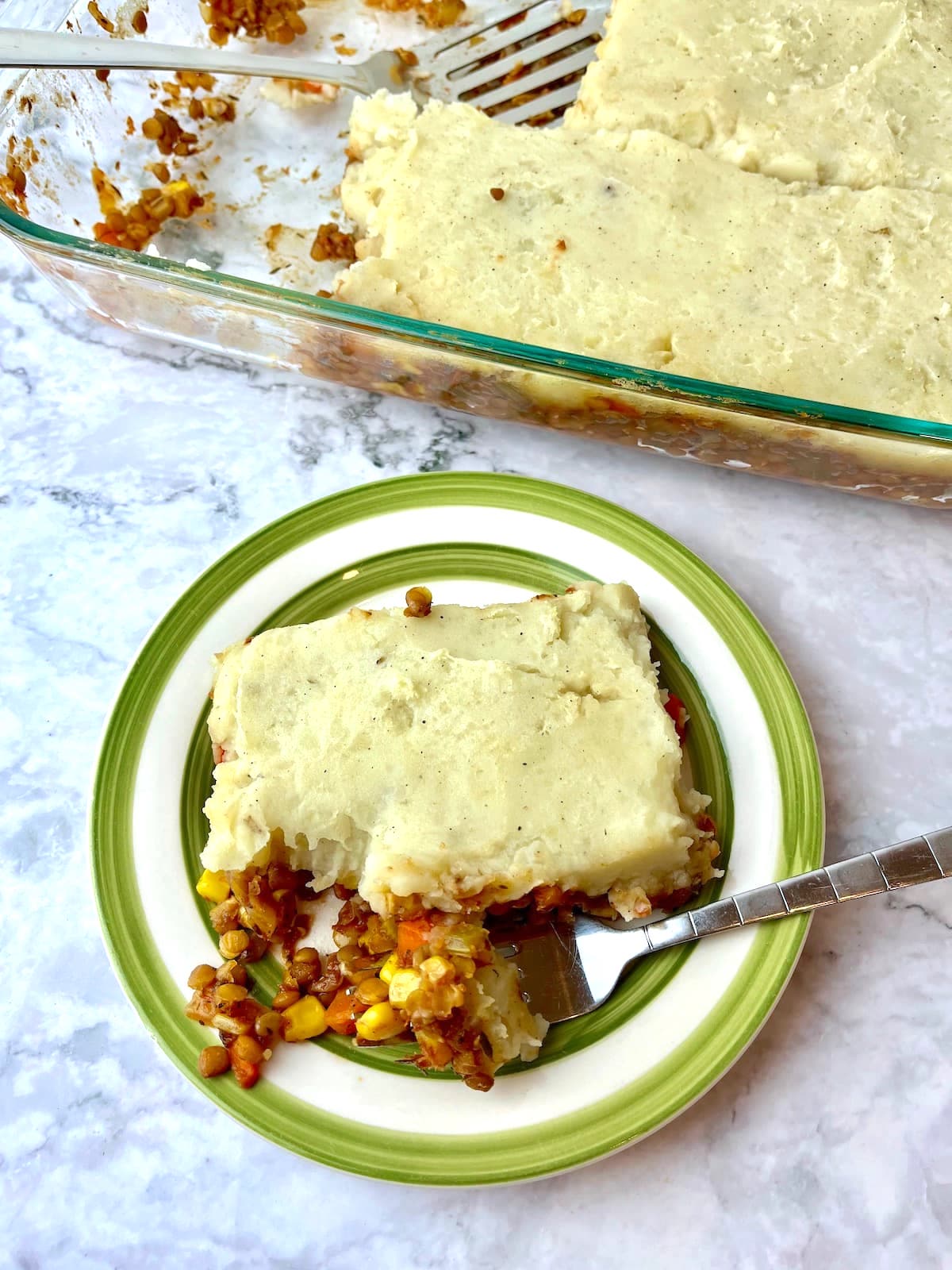 A piece of lentil shepherd's pie with a fork on a plate, next to a baking dish of lentil shepherd's pie.