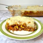 A piece of lentil shepherd's pie on a plate in front of a baking dish of lentil shepherd's pie.