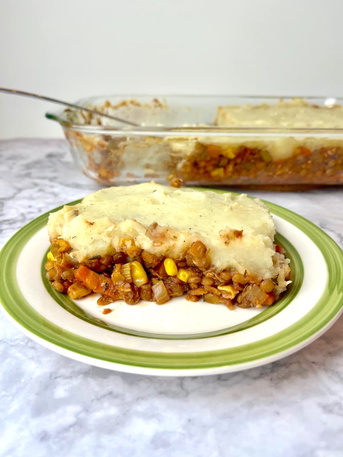 A piece of lentil shepherd's pie on a plate in front of a baking dish of lentil shepherd's pie.