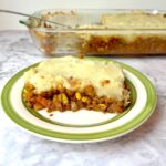 A piece of lentil shepherd's pie on a plate in front of a baking dish of lentil shepherd's pie.