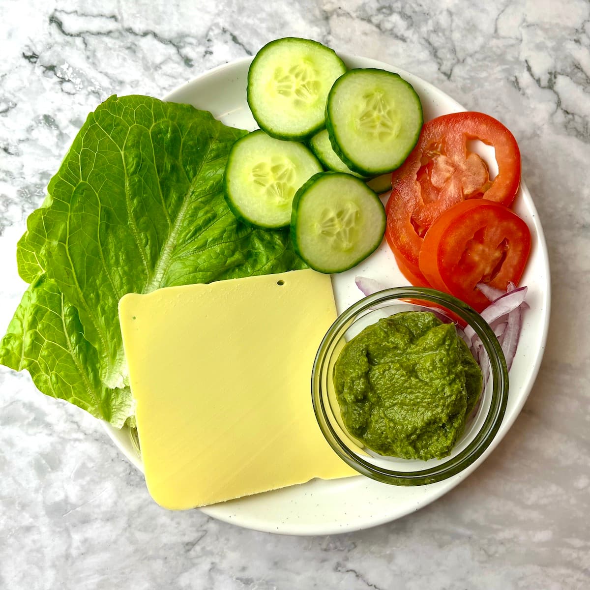 A plate of ingredients including vegan pesto, tomato, cucumber, onion, lettuce, and vegan cheese.