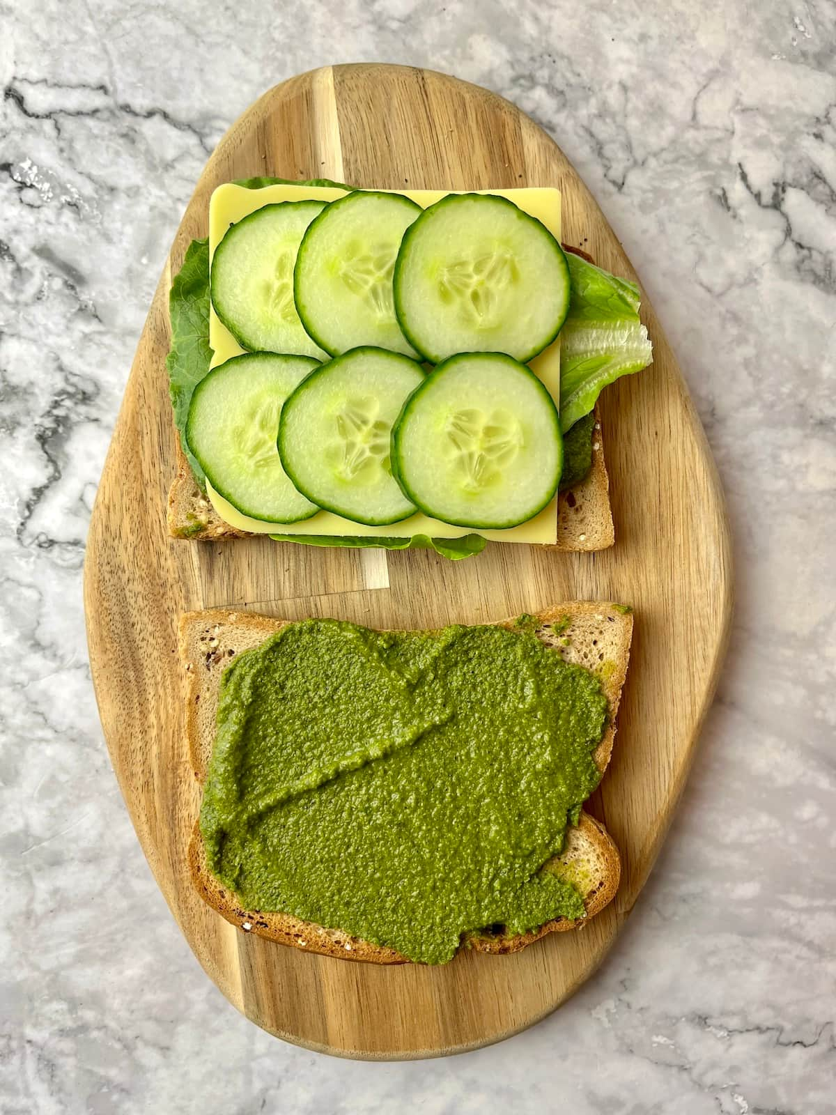 Two slices of bread, one with pesto and the other with lettuce, vegan cheese, and cucumber slices.