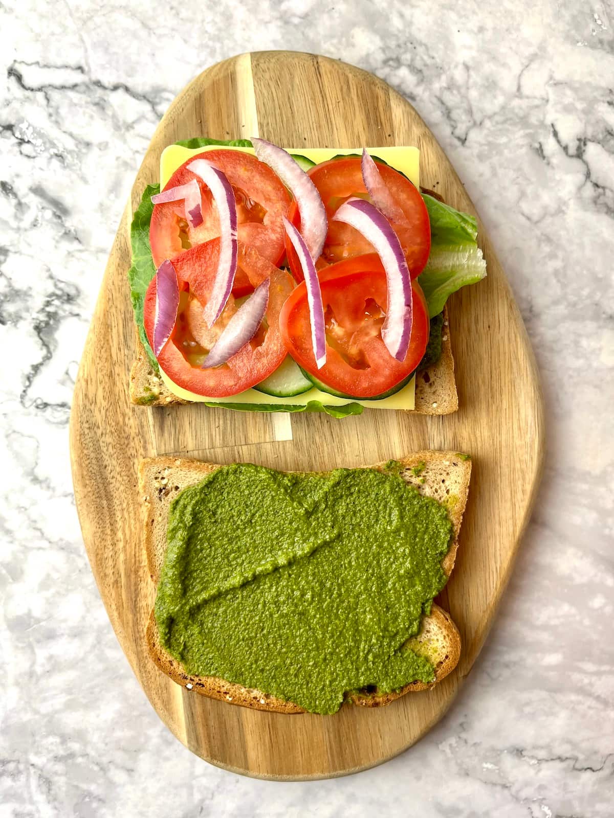 Two slices of bread, one with pesto and one with veggies including tomato and onion slices.