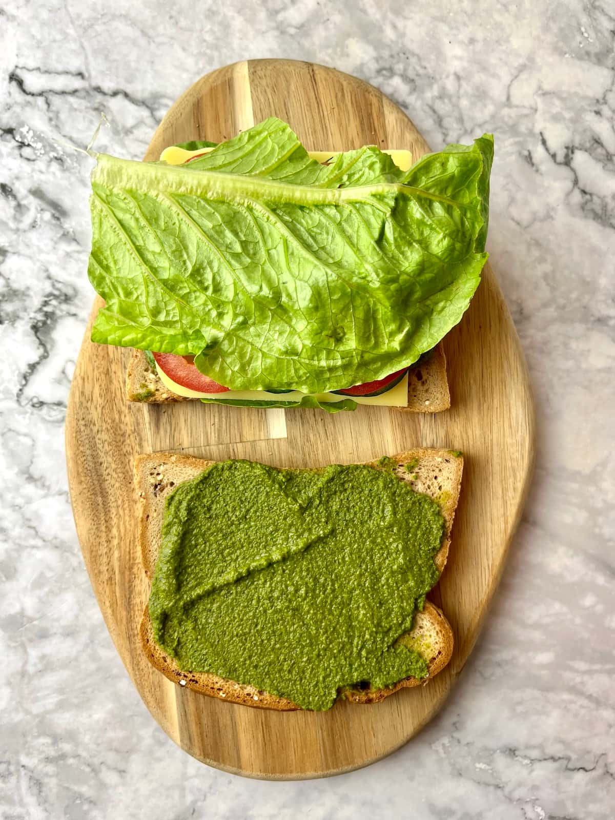 Two slices of bread, one with pesto, and one with veggies and lettuce on top.