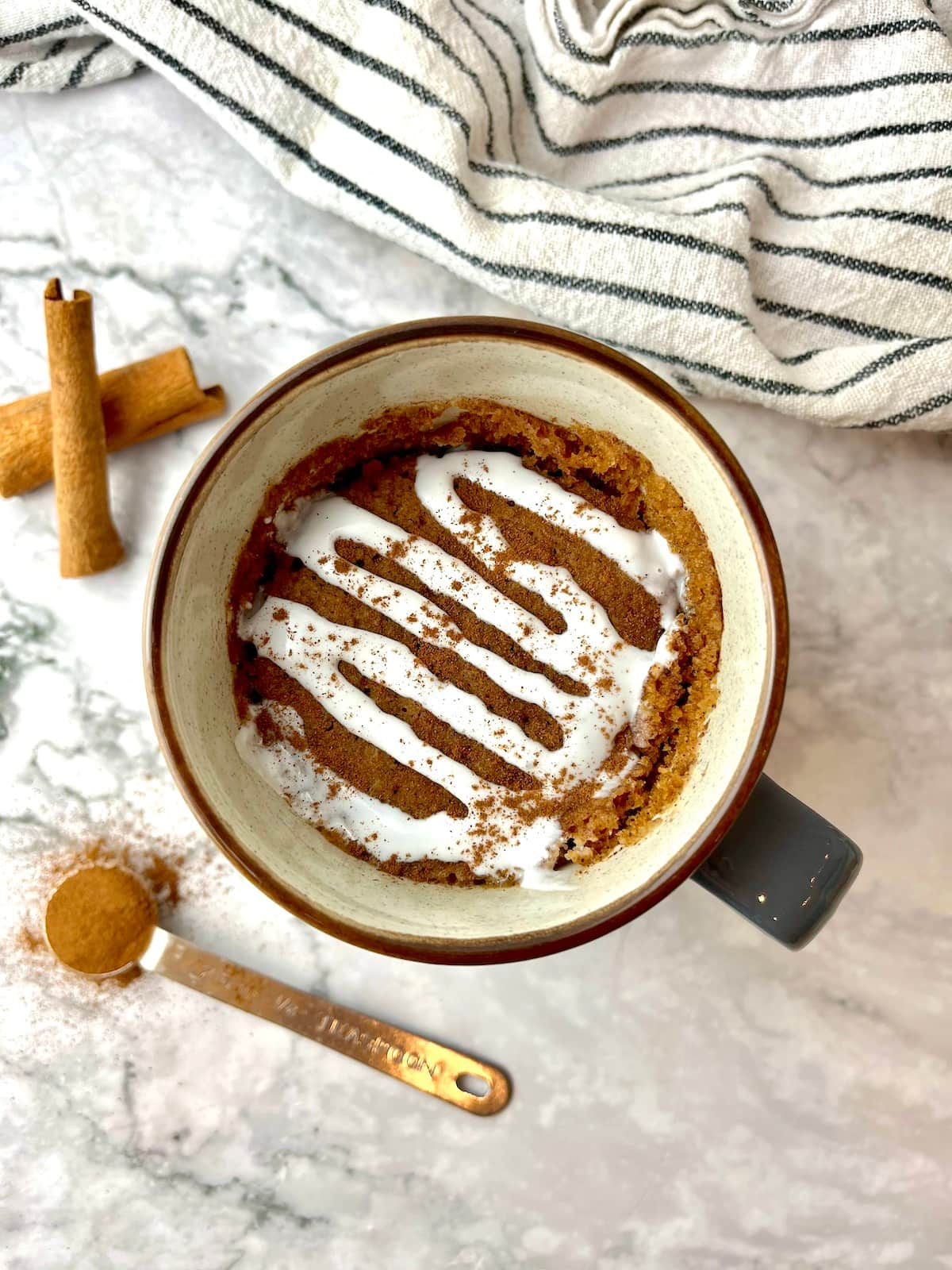 A cinnamon mug cake topped with white icing next to cinnamon sticks and ground cinnamon.