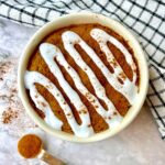 A vegan cinnamon mug cake topped with white icing and cinnamon, next to a black and white dish towel.