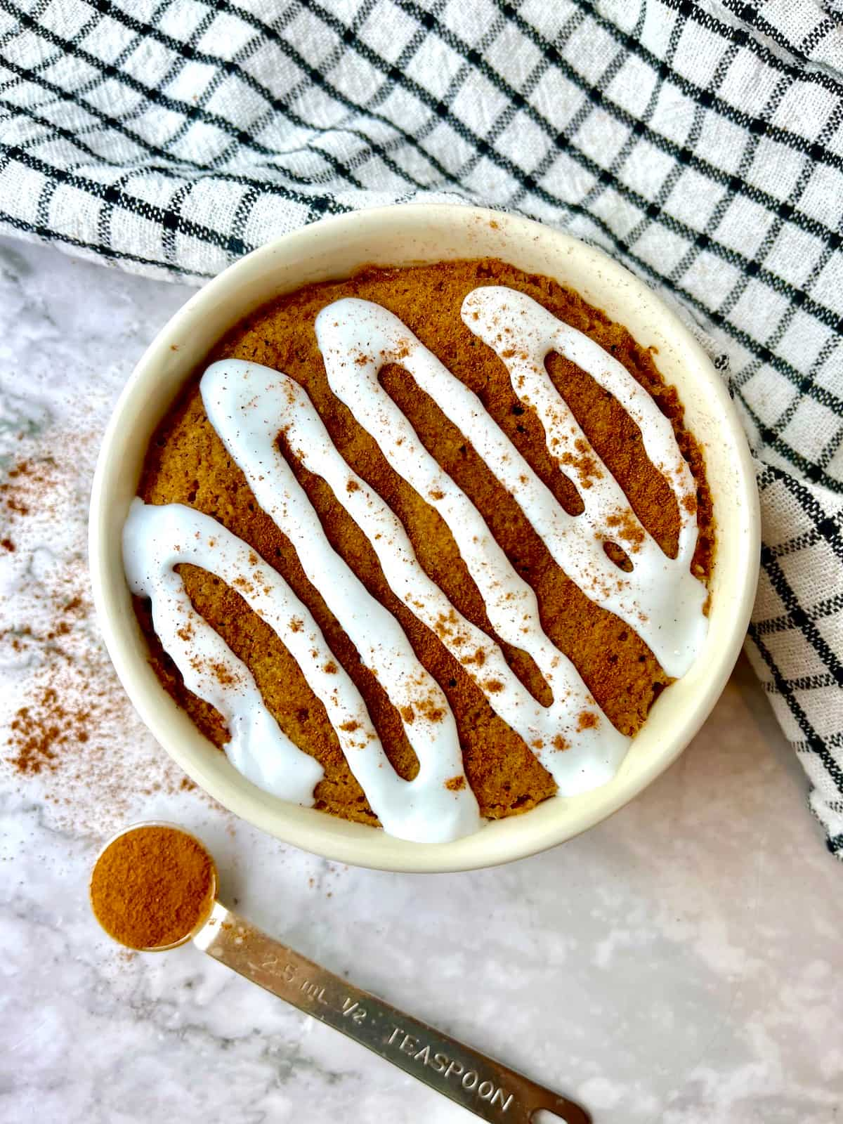 A vegan cinnamon mug cake topped with white icing and cinnamon, next to a black and white dish towel.