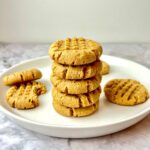 A stack of peanut butter cookies on a plate, surrounded by more peanut butter cookies.
